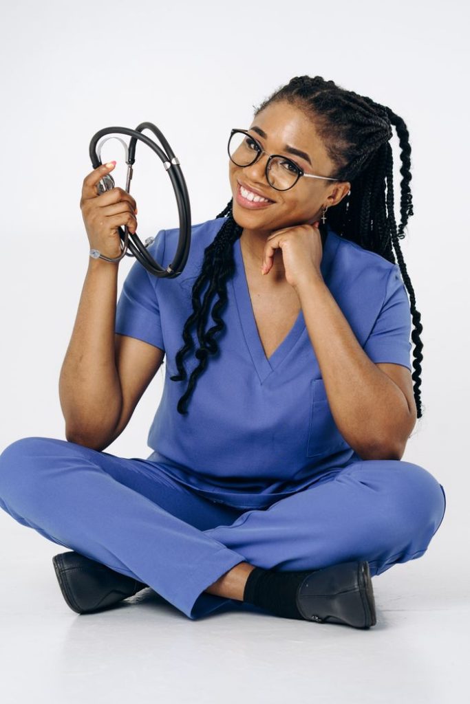 Confident female doctor in blue scrubs with stethoscope, sitting cross-legged and smiling.